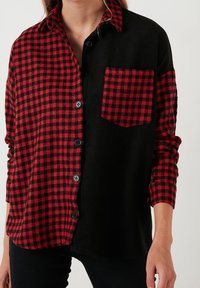 Camisa de cuadro escocés en rojo y negro con un lado y un bolsillo negros. Diseño de botones, mangas largas, textura suave y patrones contrastantes.