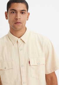 Short-sleeve beige shirt made of denim with two chest pockets, snap button closure, and a small red tag on the left pocket.