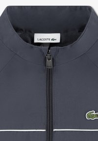 Σκούρο μπλε μπουφάν με φερμουάρ και όρθιο γιακά, με επένδυση από δίχτυ και αντίθετες λευκές ρίγες. Κεντημένο λογότυπο Lacoste στην αριστερή πλευρά.