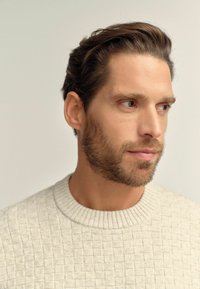 Maglione grigio chiaro a maglia testurizzata con collo a costine. In primo piano un modello maschile, con capelli corti di colore castano e una barba ben curata.