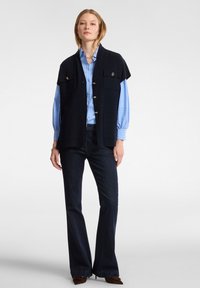 Námořnická bezrukávová vesta s akcenty knoflíků, přehozená přes světle modrou pruhovanou košili. Tmavě modré rozšířené džíny doplňují celý outfit.