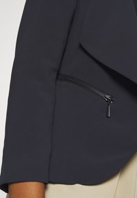 Detailní záběr na osobu v tmavě námořnické bundě s šikmou kapsou na zip a béžovými kalhotami viditelnými pod rukávem.
