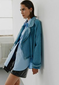 Denim jacket with a collar, paired with a blue striped shirt and a black glossy skirt. The ensemble features contrasting textures and layered styles.
