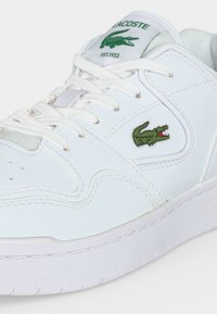 Baskets blanches Lacoste avec logo crocodile vert sur le côté et sur la languette, lacets blancs et détails de panneaux cousus.