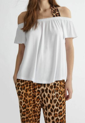 Femme portant un haut blanc à épaules dénudées avec des bretelles licou à imprimé léopard et un pantalon assorti à imprimé léopard, debout les mains dans les poches.