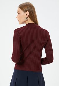 Burgunderfarbener, gerippter Pullover mit langen Ärmeln, der an den Bündchen geraffte Akzente und einen hohen Kragen hat. Glatte Textur.