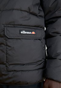 Giubbotto puffo nero con texture trapuntata, dotato di tasca frontale e logo "ellesse" ricamato. Materiale liscio, design isolato.