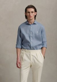 Svetlomodrá košeľa na gombíky s vyhrnutými rukávmi, v kombinácii s bielymi plisovanými nohavicami s detailmi na boku. Hladká látka a prispôsobený strih.