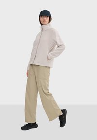 Chaqueta de forro polar beige claro con cuello alto, pantalones anchos beige y zapatillas negras texturizadas; combinada con una gorra oscura.