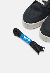 Melni Adidas kurpju šņores ar plastmasas galiem, marķētas ar zilu Adidas birku. Tuvumā ir tumšas kedas ar gaiši pelēku zoli.