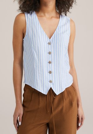 Femme portant un haut sans manches rayé bleu et blanc avec boutons et col en V, ainsi qu’un pantalon marron à cordon, sur un fond neutre.