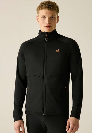 Junger Mann mit kurzen, lockigen blonden Haaren trägt eine schwarze Jacke mit Reißverschluss, orangefarbenem Logo und Besatz, steht vor einem einfarbigen beigen Hintergrund.