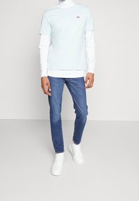 T-shirt de manga corta azul claro superpuesto a una camisa blanca de manga larga, combinado con jeans ajustados azules y zapatillas blancas.