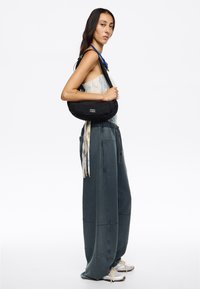 Czarna, okrągła torba na ramię wykonana z miękkiego materiału, z logo. Modelka ma na sobie luźne, szare spodnie i jasną bluzkę.