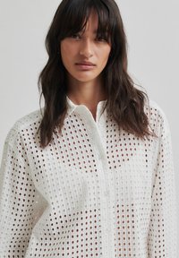 Bela srajca z gumbi, ki vsebuje vzorčasto mrežno zasnovo z majhnimi kvadratnimi izrezi, lahek material in klasičen ovratnik.