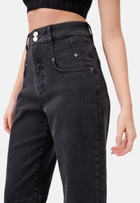 Pantaloni alti in denim nero con due tasche frontali, chiusura con cerniera e bottone, e hardware argentato in rilievo. Tessuto morbido, vestibilità dritta.