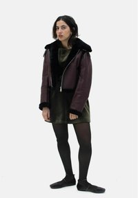 Burgunderlederjacke im Crop-Stil mit schwarzem Kunstfellkragen und -manschetten, getragen über einem grünen Samtkleid, kombiniert mit schwarzen Strumpfhosen und Ballerinas.