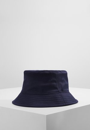 Marineblauwe katoenen bucket hat met gestikte rand en ventilatiegaatjes, tentoongesteld op een wit voetstuk tegen een lichtgrijze achtergrond.