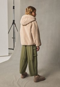 Jachetă cu glugă din fleece, de culoare bej deschis, combinată cu pantaloni largi cargo de culoare olive. Modelul poartă pantofi maro slip-on cu suprafață texturată.