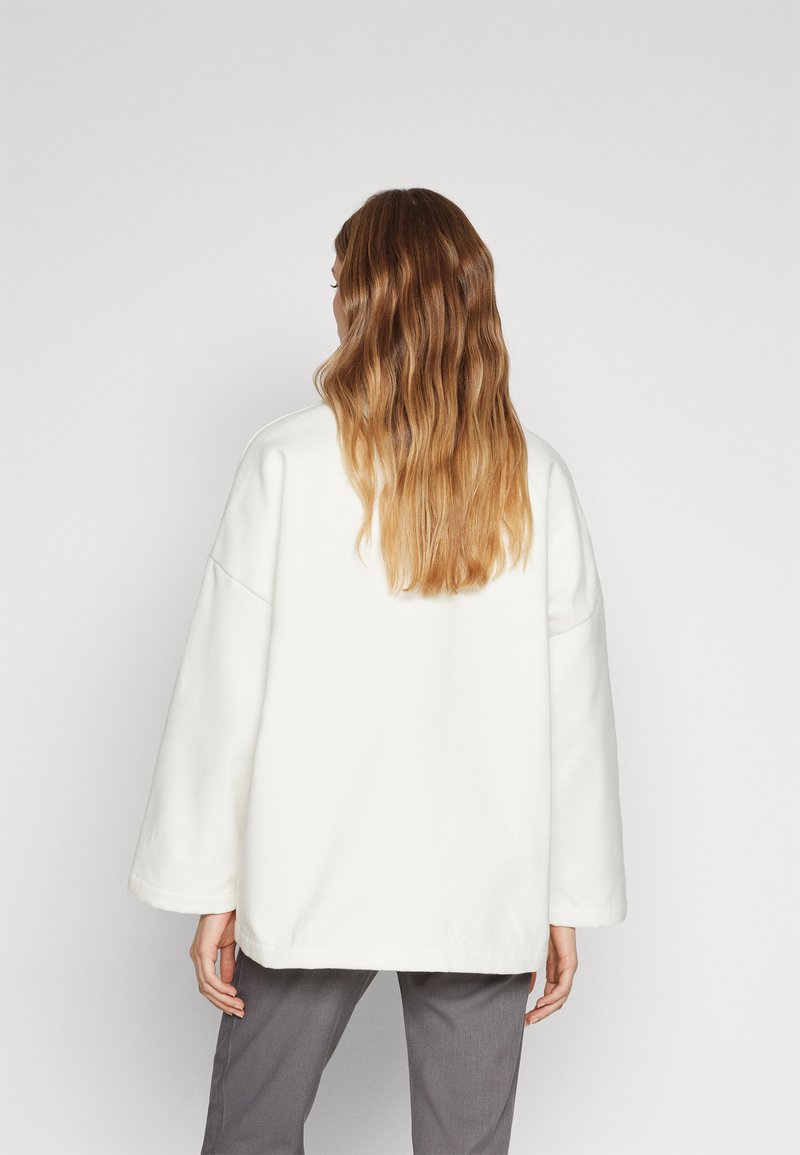 Sudadera blanca de gran tamaño hecha de material suave, con mangas anchas, corte recto y diseño minimalista. Combinada con pantalones grises.