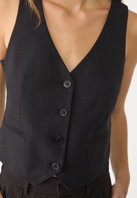 Schwarze Weste mit tiefem V-Ausschnitt, strukturiertem Design, fünf Knöpfen und Seitentaschen. Hergestellt aus strukturiertem Stoff.