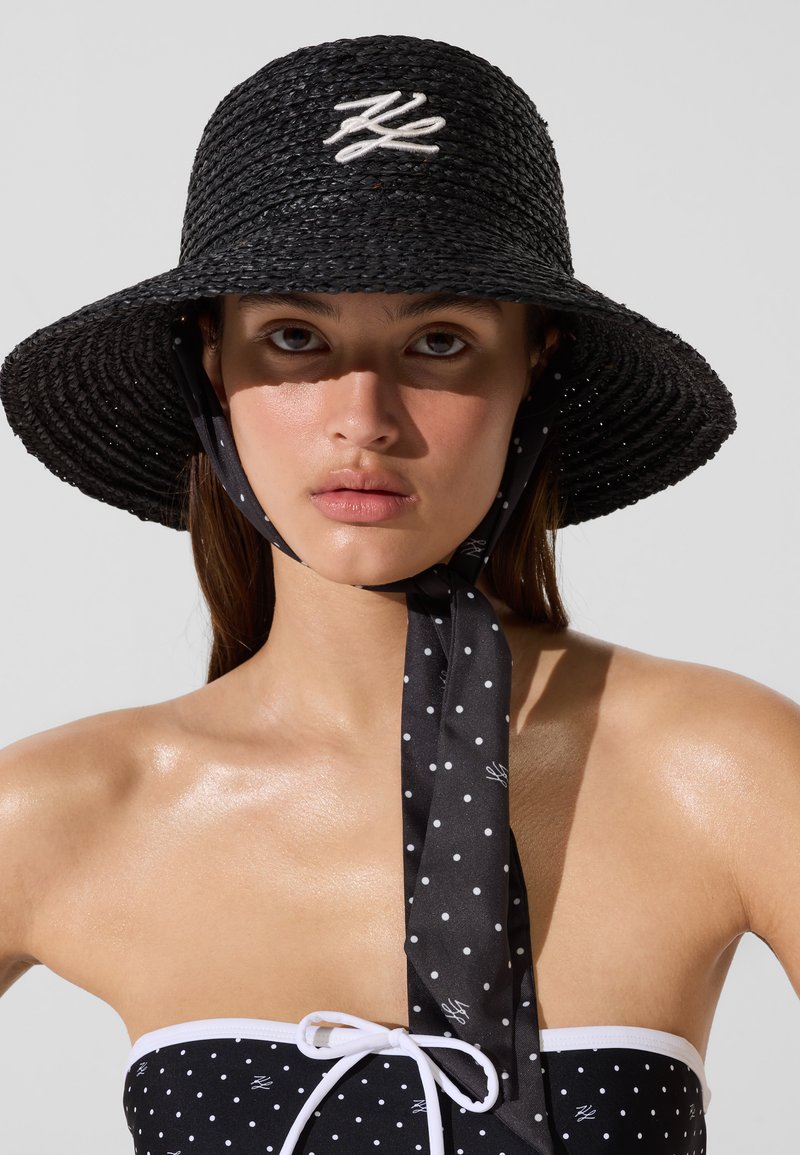 Jeune femme portant un chapeau noir à larges bords avec un lien sous le menton et un haut noir sans bretelles à pois avec une bordure blanche et un nœud.