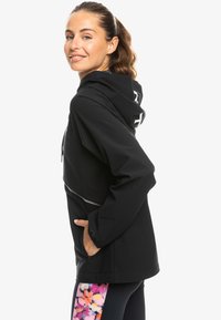 Black waterproof jacket with a hood, featuring a silver zipper pocket and a logo. Model wears leggings with a colorful floral pattern.