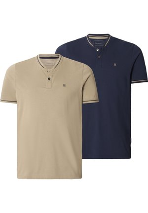 Mann, der ein beiges Henley-Shirt mit kurzen Ärmeln, zwei Knöpfen und dunklem Besatz an Kragen und Ärmeln trägt, vor weißem Hintergrund stehend.