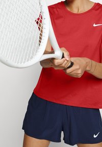 Punainen kosteutta siirtävä lyhythihainen urheilupaita, jossa on pyöreä kaula-aukko, yhdistettynä tummansinisiin shortsihin. Kädessä valkoinen tennismaila, jossa on teksturoitu pito.