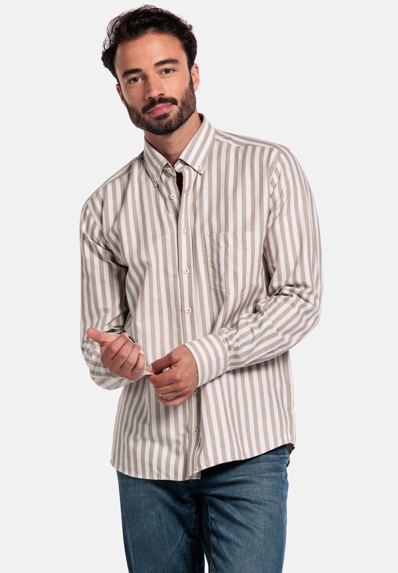 Man met een baard die een beige en witte verticaal gestreepte button-up shirt en blauwe jeans draagt, terwijl hij zijn manchet aanpast tegen een plain witte achtergrond.