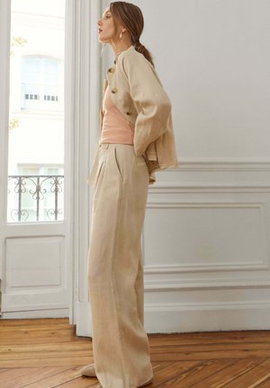 Vrouw staat binnen en draagt een beige jas draperend over haar schouders, beige wijde broekspijpen en een perzikkleurige mouwloze top.