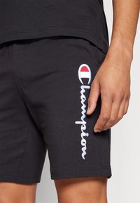 Shorts de algodón negros con logo "Champion" bordado en blanco y emblema colorido. Primer plano de la textura del tejido y detalles de las costuras.