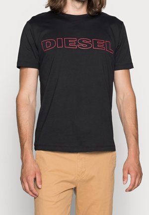 Homme portant un T-shirt noir à manches courtes avec le texte "DIESEL" en rouge contour sur la poitrine, et un pantalon beige, cadré au niveau du cou et à mi-cuisse.