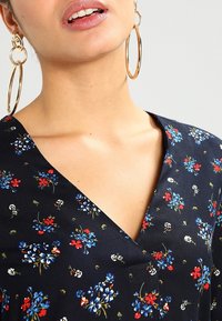 Květinové šaty s námořnickým pozadím, zdobené červenými a modrými květinami. Měkký výstřih do V, hladká textura, doplněné velkými zlatými kroužkovými náušnicemi.