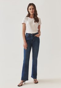 Jeans de mezclilla azul de tiro alto y pernera acampanada con un acabado ligeramente desgastado, combinados con una camiseta blanca de manga corta y chanclas negras.