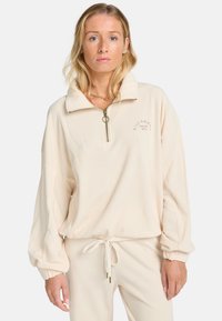 Ecrufarbener, gerippter Pullover mit einem Reißverschlusskragen, langen, gepufften Ärmeln und einem Kordelzugsaum. Mit dezenter Logo-Stickerei auf der Brust.