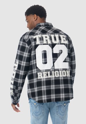 Mann trägt ein schwarz-weiß kariertes Hemd mit großem weißem Text "TRUE 02 RELIGION" auf Rücken und Ärmel, kombiniert mit hellblauen Jeans.