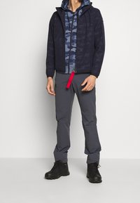 Herreudendørsoutfit bestående af en navy mønstret jakke, en blå camouflage hættetrøje med rød lynlås, grå bukser og sorte støvler.