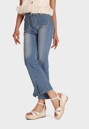 Jeans in denim blu chiaro con cuciture floreali decorative sulle tasche, leggermente svasati all'orlo e con fessure laterali. Accoppiati con sandali con zeppa a strascico beige.