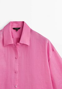 Chemise rose pâle boutonnée avec col évasé, manches courtes et texture légèrement transparente, dotée de boutons assortis rose sur le devant.