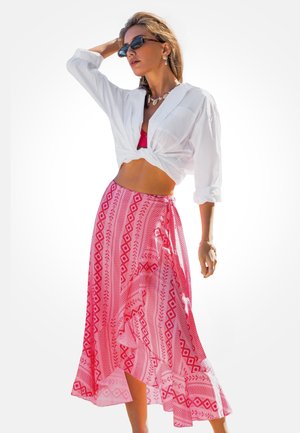 Weißes, geknotetes Hemd, roter Rock mit geometrischen Mustern. Der Rock ist aus einem fließenden, leichten Stoff. Accessoires umfassen eine dunkle Sonnenbrille.