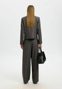 Harmaa räätälöity puku, jossa leveälahkeiset housut ja lyhyt bleiseri. Pitelee pientä mustaa käsilaukkua, jossa on solmupainatus. Siisti, jäsennelty muotoilu.