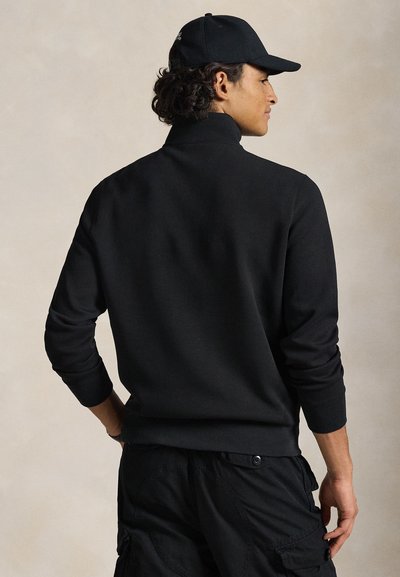 Svart genser med høy hals, lange ermer, ribbede mansjetter og glatt tekstur, kombinert med svarte cargobukser og en cap.
