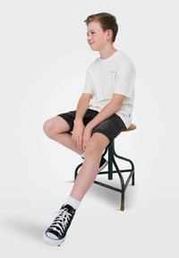 Vit bomulls-t-shirt, grå denimshorts, svarta högskurna sneakers med vita sulor, sitter på en grön metallpall framför en ljus bakgrund.