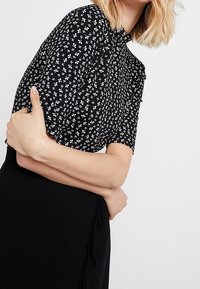 Blusa negra con pequeños patrones florales blancos, mangas cortas y cuello con solapa. La tela parece ligera y texturizada.