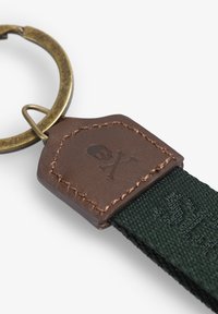 Llavero con un anillo de bronce y una correa de tela verde, que cuenta con una etiqueta de cuero marrón texturizada grabada con un motivo de calavera y huesos cruzados.