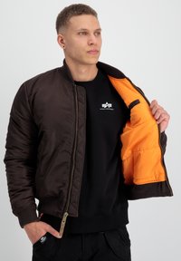 Jachetă bomber maro cu textură matlasată, căptușeală portocalie, manșete cu nervuri și închidere cu fermoar. Asortată cu un hanorac negru dedesubt.