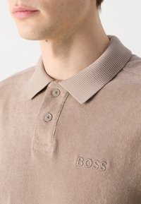 Gros plan d'une personne portant un polo beige avec col côtelé, deux boutons et "BOSS" brodé sur la poitrine.