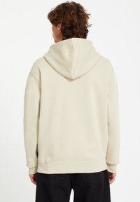 Beige Kapuzenpullover mit weicher Textur, gerippten Bündchen und Saum. Verfügt über eine Kängurutasche und wird von hinten gezeigt.