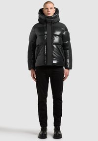 Doudoune noire avec col haut, finition brillante, fermeture éclair et deux poches avant. Associée à un pantalon noir et des bottes.